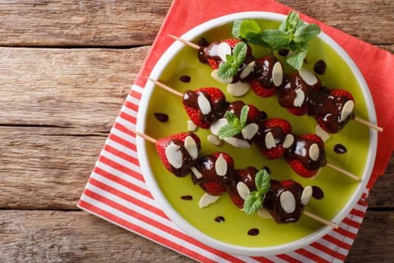 Strawberries on fruit skewers topped with chocolate sauce, almond slices, and mint leaves, served on a white plate with a green base on a red striped napkin—a delightful chocolate dipped fruit treat.