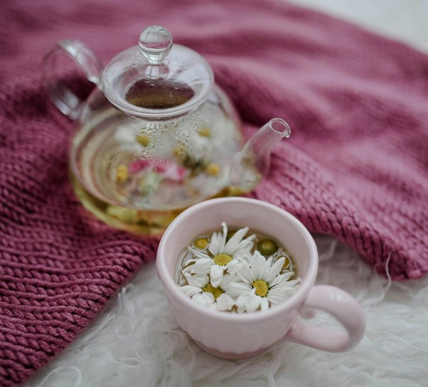 A glass teapot and a pink cup with soothing Chamomile Tea sit on a white surface next to a folded mauve knitted blanket—a perfect bedtime soother.
