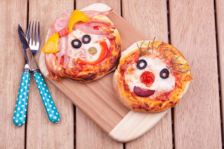 Two mini pizzas decorated as playful pizza faces with olives, peppers, and herbs, arranged on a wooden board with a fork and knife—perfect for a fun DIY meal.