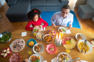 A man and a girl sit at a dining table set with various dishes, drinks, and lit candles, preparing to eat—perfect for introducing variety to kids’ meals or finding meal ideas for selective eaters.