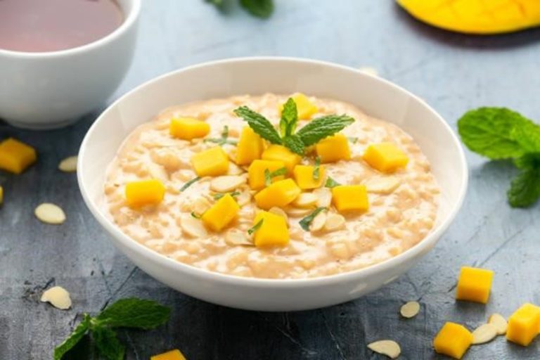 A bowl of creamy coconut rice pudding topped with diced mango and mint leaves, with almond slices and a cup of tea in the background.
