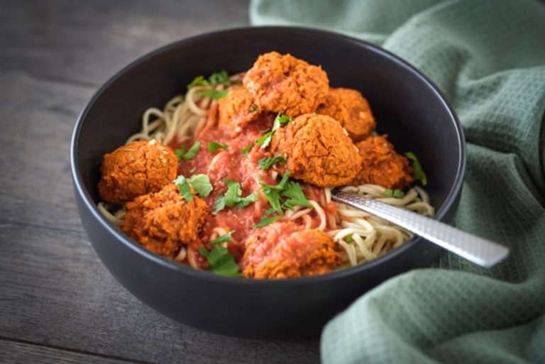 A black bowl filled with spaghetti bolognese, topped with tomato sauce, several meatballs, and garnished with chopped parsley, with a fork resting inside the bowl.