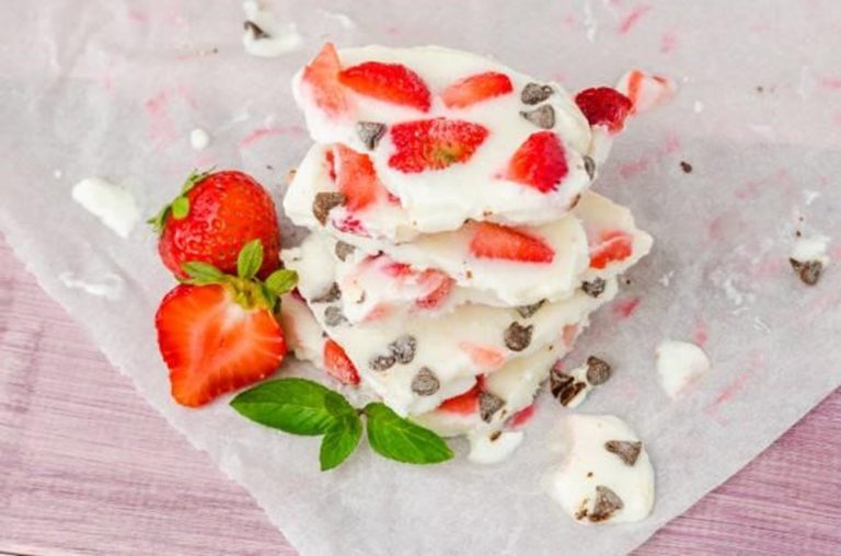 Stack of Strawberry Yogurt Bark pieces with strawberry slices and chocolate chips on parchment paper, next to fresh strawberries and a green mint leaf.