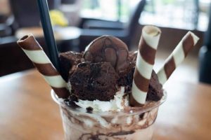 A Bliss Shake dessert cup with chocolate ice cream, whipped cream, brownie pieces, chocolate sauce, and three chocolate wafer rolls, served with a black straw. Perfect for fans of rich Chocolate Brownie flavors.